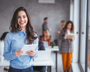 Was ist Corporate Learning? Eine lächelnde Frau in einem blauen Hemd hält ein Tablet in der Hand, während sie in einem modernen Büro steht. Im Hintergrund sind mehrere Personen bei der Arbeit zu sehen.